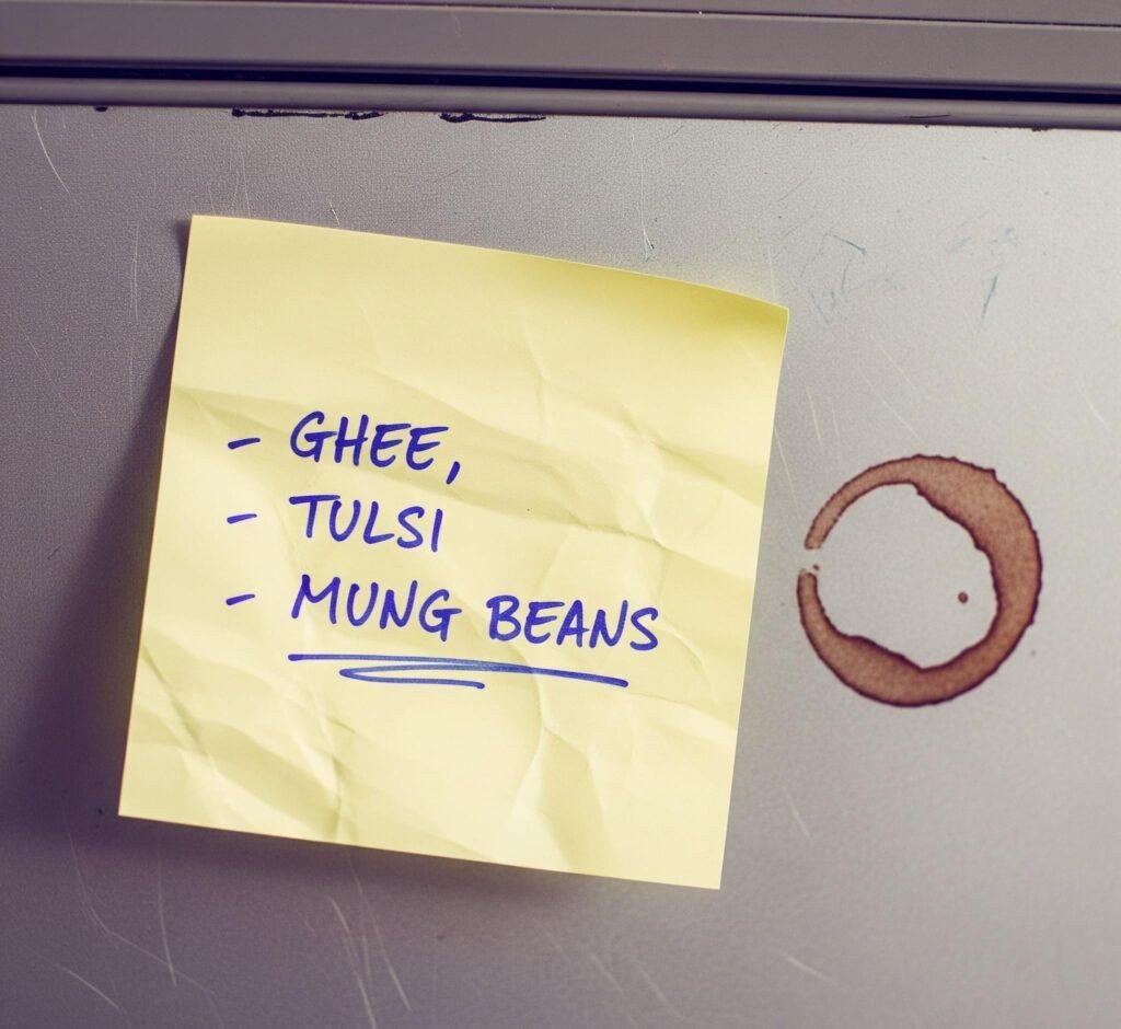 A crumpled sticky note listing ingredients on a fridge, with a coffee ring nearby.